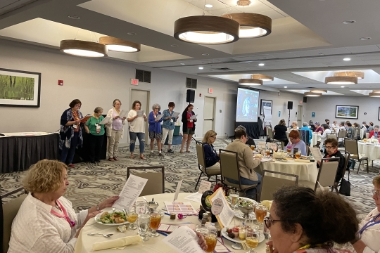 women sitting around tables and also standing in a room. 