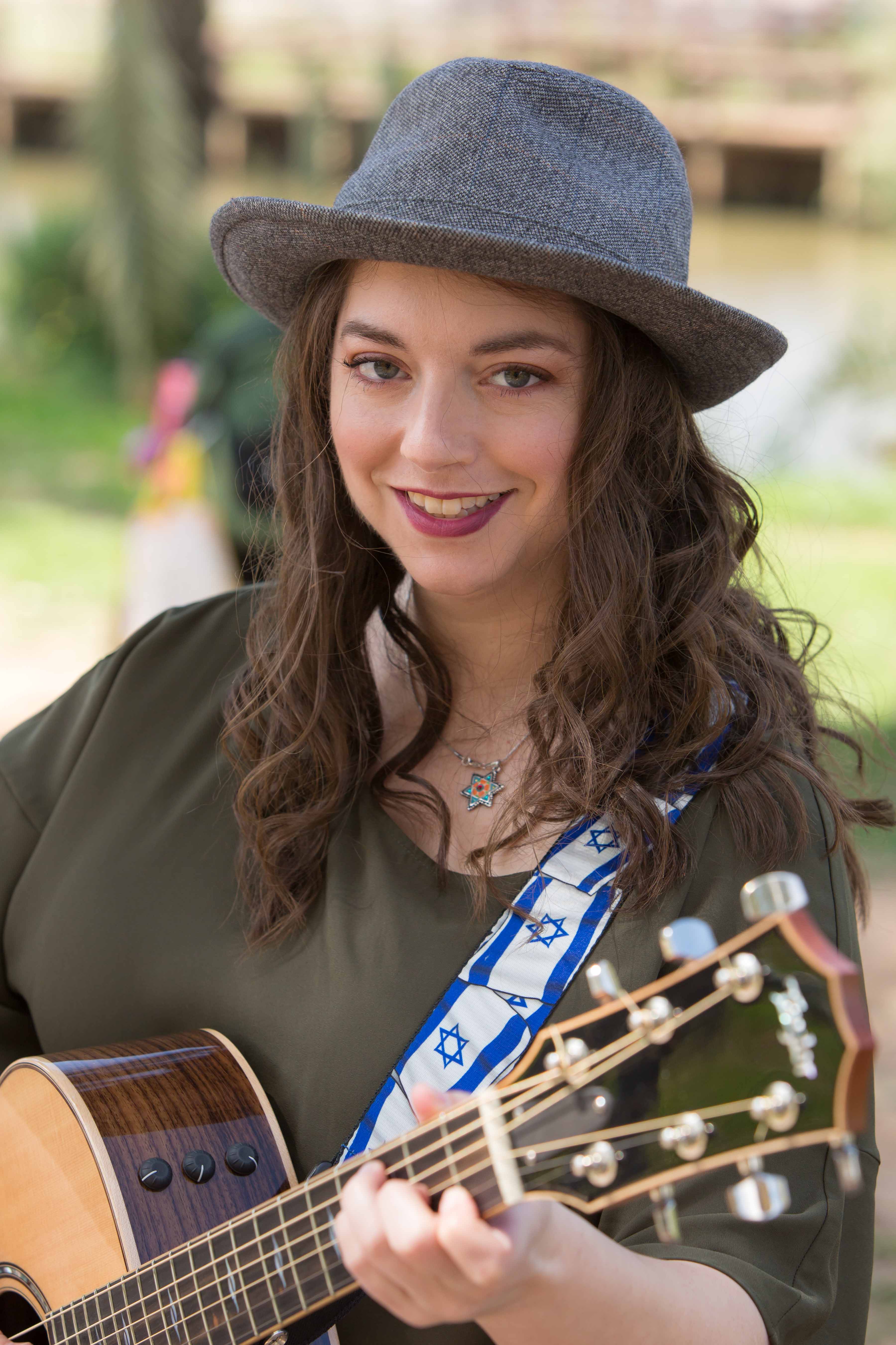 A woman with brown shoulder length hair, gray hat, green shirt, red lipstick, smiles and holds a guitar. 