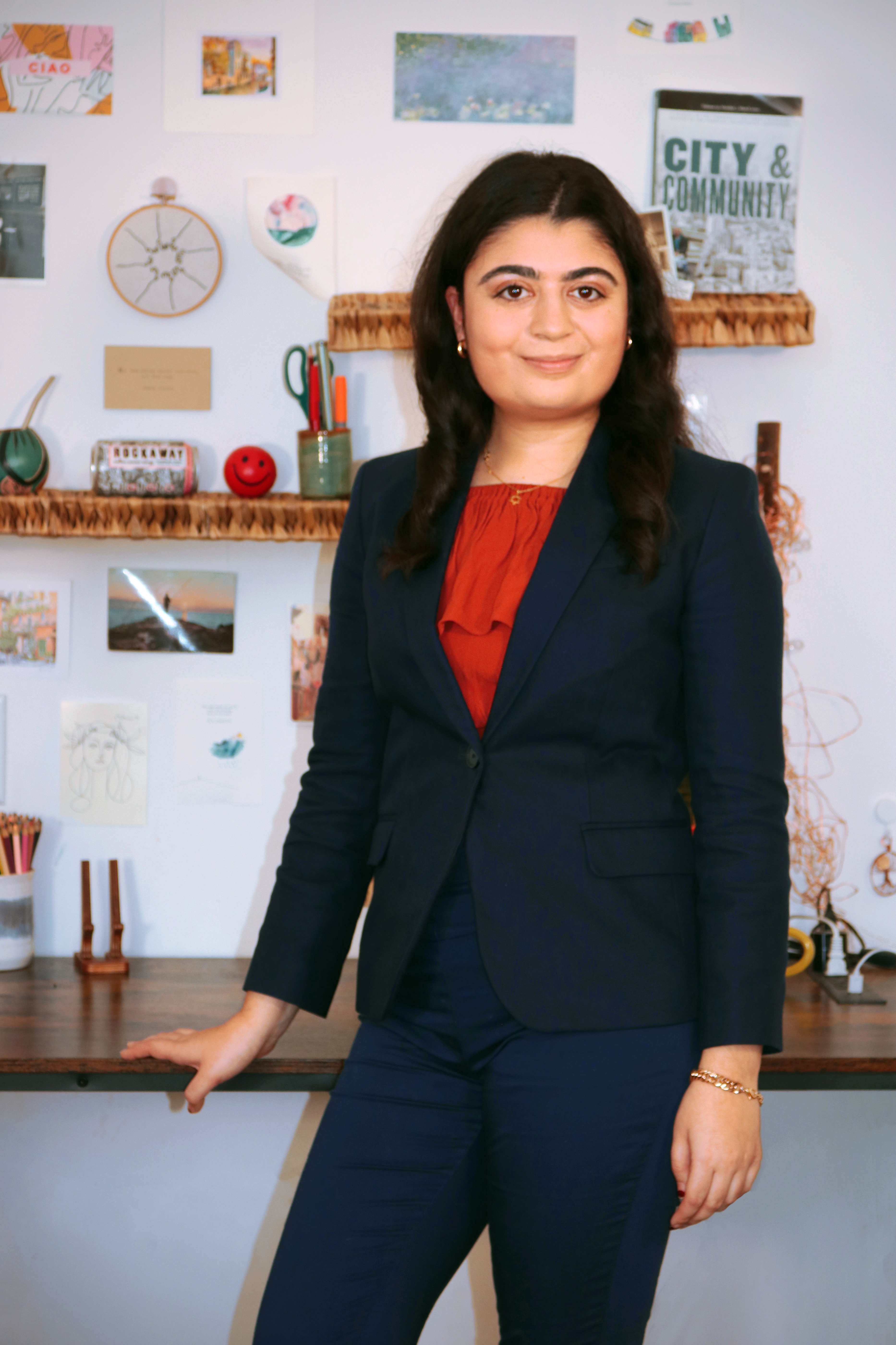 A woman smiling with shoulder length brown hair in a blazer and red shirt stands in front of a desk. 