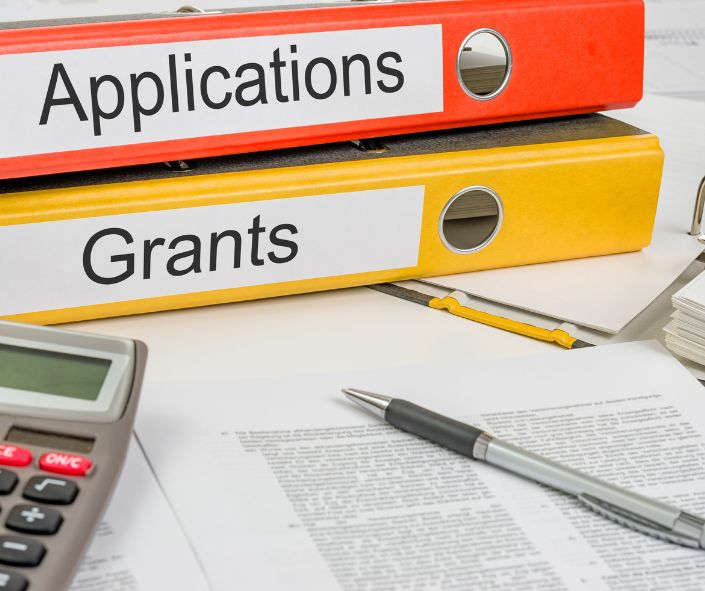 Red and yellow binders with "Applications" and "Grants" printed, a calculator, pen, and some documents. 