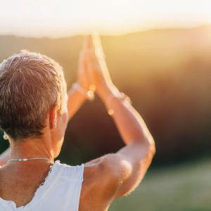 A woman in a white tank top with short gray hair hold her hands up in prayer position while facing a sunny sky and blurred green mountains/hill. 