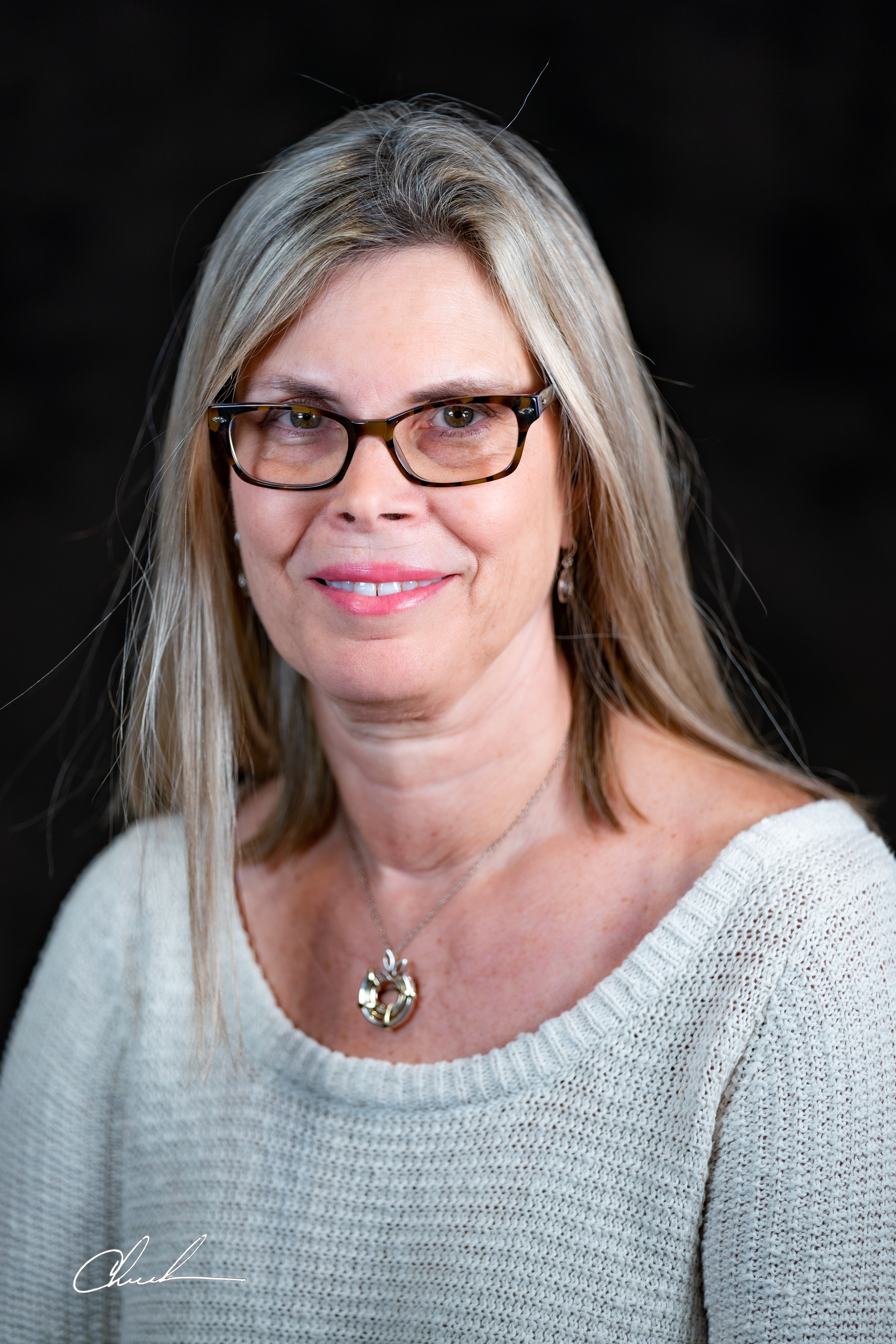 A woman with shoulder length blond hair and dark square frames, wearing a white long sleeve top, and a circular metal necklace smiles against a black background.  